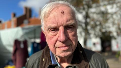 Brian Burbidge frowns as he stands in a market place and looks into the camera. He has short white hair and is wearing a green and brown wax jacket.