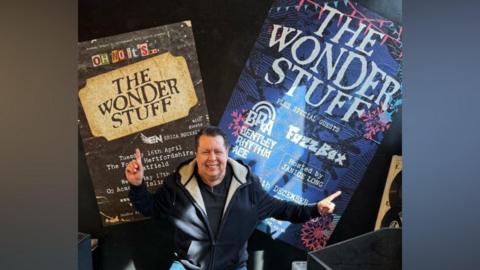 Middle aged man with a hoody on pointing behind him to The Wonder Stuff band posters