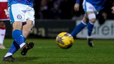 Two Stockport County football players can be seen from the torso down playing on a field. The man in the foreground has just kicked the football. 