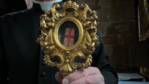Close up image of vicar's hand. The vicar is wearing balck clothing with a white collar and is holding an intricate gold painted reliquary which houses a small wooden box which is tied by a red ribbon making the shape of a red cross.