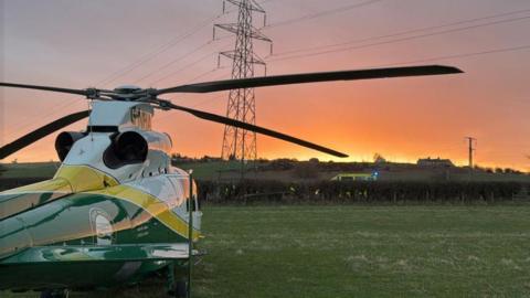 A white, green and yellow air ambulance stationary in a field. In the background an ambulance can be seen behind a hedge. There is a pylon and the sky is orange and yellow. 