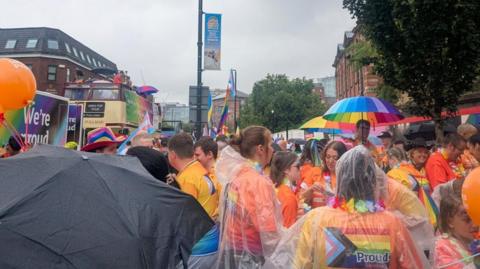 A group of people, mostly clad in orange, huddled on city centre street. Some of them are wearing waterproof ponchos while others are holding umbrellas. An open top double decker bus is in the background.