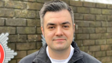 A man with salt and pepper short grey hair who is wearing a black coat stands in front of a brick wall with a fire and rescue logo on it.