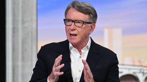 Close-up of Peter Mandelson, wearing a black jacket and white button-up shirt and black spectacles, speaking at a BBC studio.