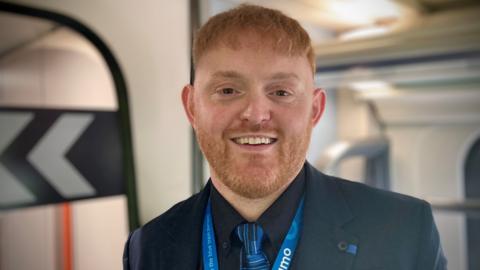 Close-up picture of Liam Dainty standing in a railway carriage wearing his uniform of a dark coloured jacket and shirt with light blue tie and Lumo lanyard. He has short ginger hair and beard.