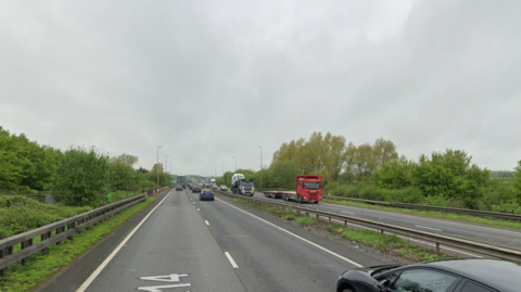 A Google view of a dual-carriageway, with multiple cars and lorries. On both sides of the road are trees and street lights.