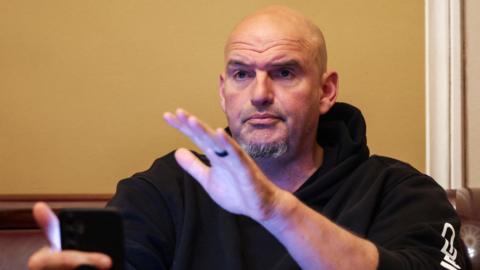 John Fetterman raising his left hand, wearing a black hoodie