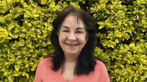 Cheryl Kenealy is a woman with dark hair and dark eyebrows, wearing a pink jumper. She is standing in front of a green bush and smiling into the camera.