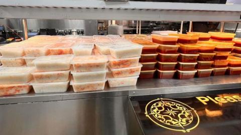 Dozens and dozens of tuppaware are stacked on a metal counter. The left half are filled with rice, the right half are filled with a red curry.