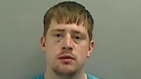 Police custody image of Jamie Govan. He has brown hair brushed down over his forehead and is wearing a light blue top. He has cuts and bruising on his face.