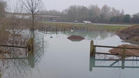 A field with a flooded section in the middle where a burst pipe has overflowed the basin. The floodwater is surrounded by wooden and metal fencing. In the background you can see cars driving along the A419. It is a grey and misty day.