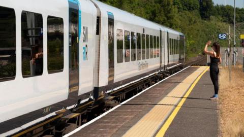 A woman waving from platform as a white South Western Railway passenger train departs Micheldever Station.