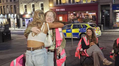 Two of the volunteers in pink hi-vis jackets. The woman on the left has ginger hair tied back and is standing up and hugging a woman with blonde, long curly hair while smiling. The volunteer on the right is sitting down in a camping chair and has brown long hair and is also smiling. There is a police car parked behind them and a row of restaurants and shops behind that.
