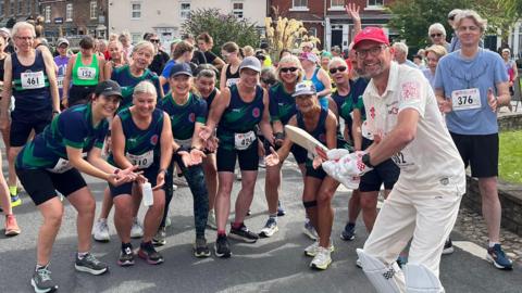 A man wearing cricket whites is standing in the middle of the photo. He is wearing gloves, shin pads, and is holding a cricket bat as if to strike the ball. He has glasses and a red baseball cap on. He is surrounded by runners pretending to be ready to catch the ball.