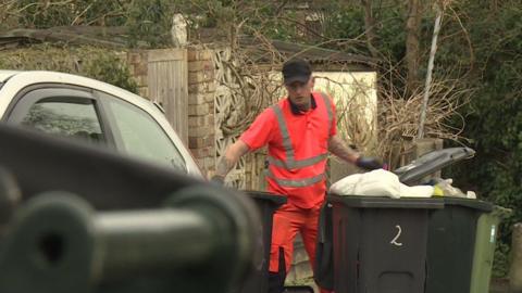 A man wearing pink high visibility clothing. He is wearing black gloves and a black cap. There are three black bins around him. 