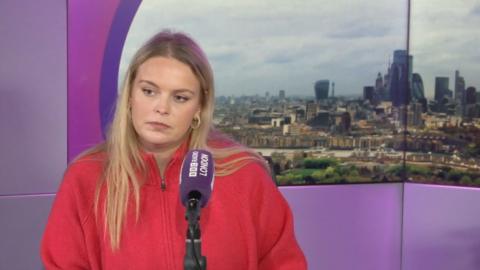A woman in a red sweater stands in front of a cityscape backdrop behind a microphone with "BBC Radio London" branding.