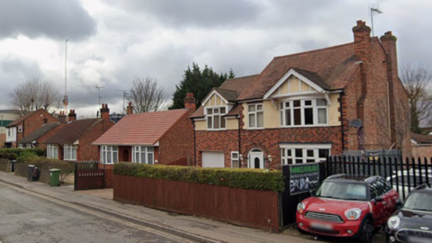 A row of detached houses, two of which appear to be bungalows. They are next to a car dealership, with two Minis parked out the front by a black fence.