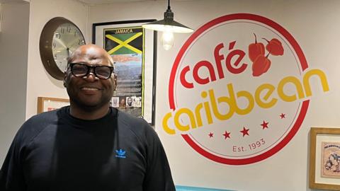 Warren Richards, who has a shaved head and is wearing black glasses and a black Adidas jumper. He is standing inside his restaurant, which has various framed items inspired by Jamaica on the wall, including its green, yellow and black flag. Also on the wall are the words "Café Caribbean".