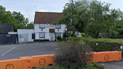 A Google Maps picture of the pub, a two-storey building with white render on it with windows and doors that have been covered by metal covers. There is a large orange barrier outside the building and a bush or hedge just in front of it.