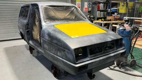 Grey body of a three-wheeled car, with yellow rectangle on the bonnet. There are sheets covering the interior. The headlights and other furniture from the front are missing. The car is inside a workshop with a roller shutter.  There is a blue canister to the right.