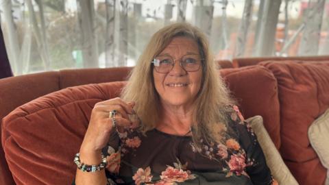 A women with dark blonde mid length hair smiling at the camera. She is wearing large glasses on her face. She is wearing a black floral top. You can see her right hand which has silver rings on with dark stones. She is also wearing a pendent bracelet.