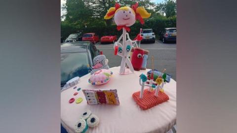 A post box topper with a large knitted girl who is shaped like a pin with scissors for legs. She has two blonde bunches and red ribbons in them. Around her are knitted sewing items, including spools of thread, smaller pins, buttons, a tape measure and a pin cushion.