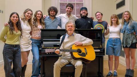 Sir Paul McCartney sits in font of a black piano and is holding an acoustic guitar. He is surrounded by nine students who all have their arms around one another.