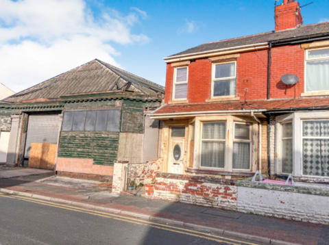 A wide shot of the front of the house. It has a white door, bay windows and orange brickwork. The front wall is discoloured, and the gate is open.