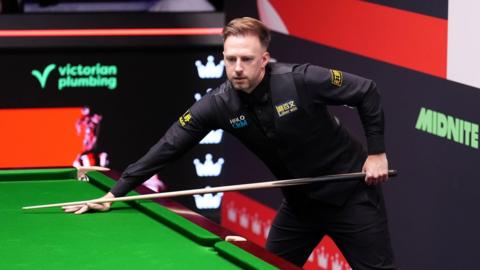 Judd Trump stands by the snooker table with his left arm holding the back of a cue and right arm on the table with cue between his fingers