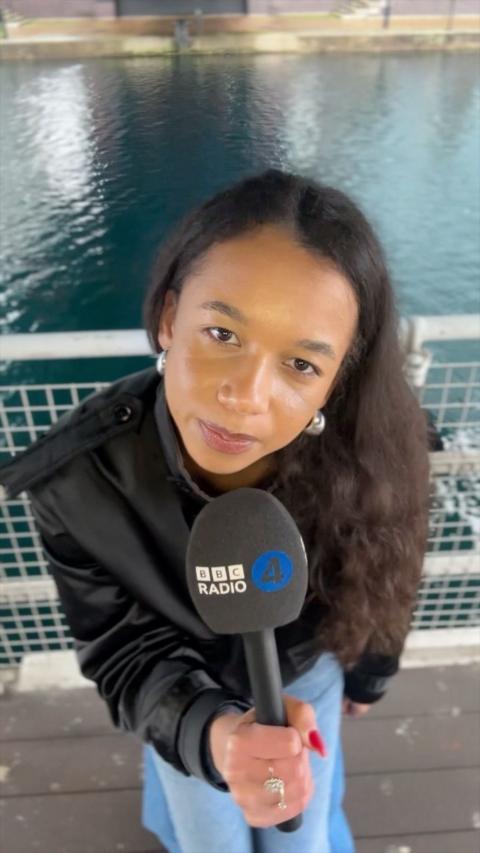 A woman with long dark curly hair holds a Radio 4 microphone. She is wearing blue trousers and a black jacket.