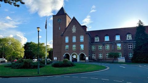 The red/brown brick headquarters of Spelthorne Borough Council, a building with slanted rooves, a clock tower and a green space and roads in front of it. There is a flagpole on the green space.
