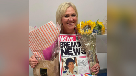 Mary Donaldson pictured with a bouquet of sunflowers, leaving gifts and a large leaving card which is designed to look like a tabloid paper. She has blonde hair, silver rings on her fingers and is smiling at the camera.