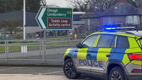 A police car parked in front of a road sign reading 'Omagh Londonderry A5'. 