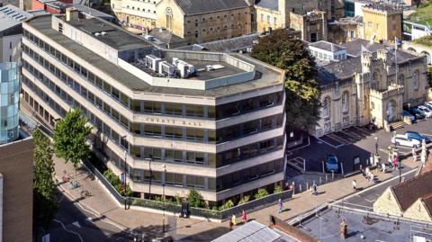 An aerial image of the current County Hall building on a sunny day. People are walking on the streets around it. To the right is the old County Hall which is castellated and currently home to the Coroner's Court