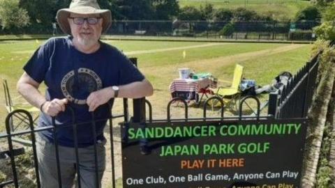 Ken Smith, a man with glasses and a white beard, wearing a hat, stands at the gate to the Sanddancer Community Japan Park Golf course, with the course behind him