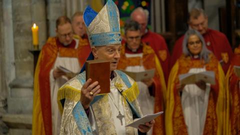 The Right Reverend Dr Mike Harrison being enthroned in Exeter Cathedral as the 72nd Bishop of Exeter. He is in official robes, which are white, blue and yellow, and is holding a bible in his right hand. Behind him is stood five men and one woman, in red, orange and white robes.