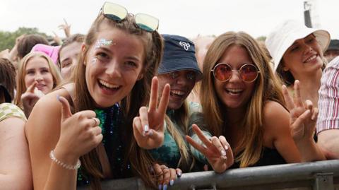 A generic outdoor festival crowd photo showing three women smiling at the camera and giving V-signs wearing sunglasses, hats and sparkly make up