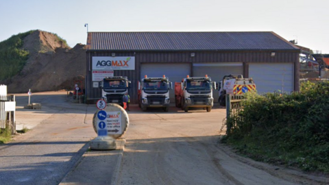 An entrance to a quarry. A building can be seen with four HGV style vehicle parked outside of it in a row. A large mound of earth can be seen to the left of the building.