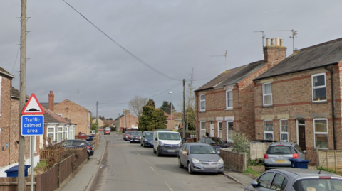 Google Street View image of Ramnoth Road in Wisbech. There are houses either side of the road and a sign on the left-hand sign of the picture that reads "Traffic calmed area". There are cars parked on the road and the sky
is a stormy grey colour.