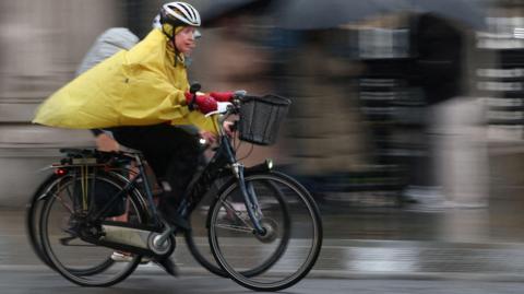 Dressed in a bike helmet and yellow rain poncho a cyclist takes to the streets, with a blurred background and another cyclist behind her. 