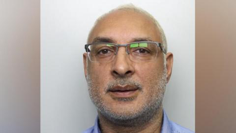 A man in his 50s, pictured in a police mugshot. He has very thin white hair, white stubble and glasses. He is wearing a blue buttoned shirt and staring directly at the camera.