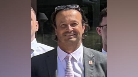A man is smiling and wearing a suit jacket over a white shirt, with a patterned tie. He has propped his glasses on his forehead.