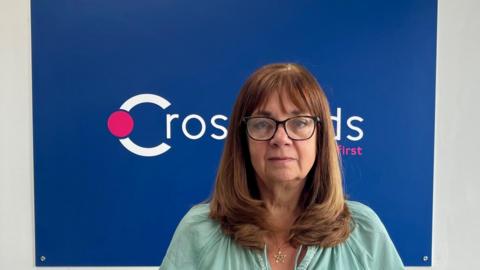 Jackie Betteridge, a woman with brown hair and a fringe, wears glasses. She is photographed in front of a predominantly blue sign that reads Crossroads.