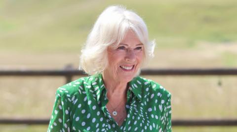 Queen Camilla is wearing a green and white polka dot blouse. She is standing outside. A grass field and wooden fence is in the background.