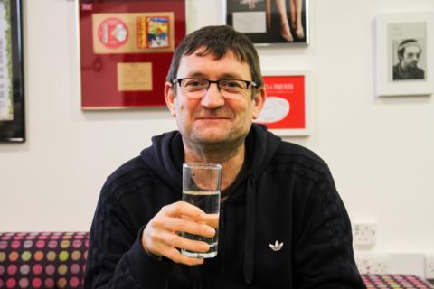 A man with brown hair and glasses sits on a sofa surrounded by pictures. He is wearing a dark hoodie jumper and is holding a glass. The wall behind him is white and covered in large colourful pictures and records.