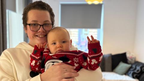 A mother with short cut hair and glasses wearing a white hoodie smiling holding her baby in a red christmas jumper. both are looking at the camera and the mother is smiling. they are in living room of a new flat.