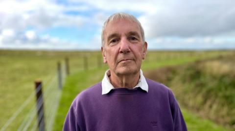 Nigel is standing on a path and you can see green fields and a blue sky in the background. There is a fence running next to him. Nigel is wearing a purple jumper and a white shirt underneath. 