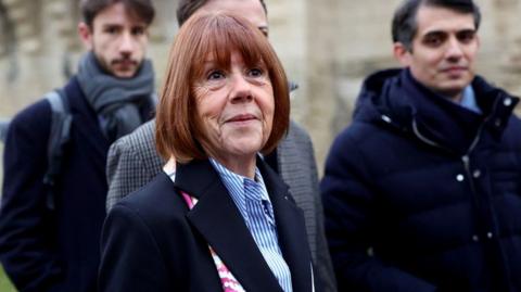 Gisèle Pelicot faces towards the camera as she arrives at court. She's wearing a dark blazer and blue and white shirt