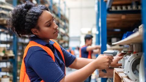 Young woman in a hi-vis vest using a digital barcode reader for her work along a shelf in a warehouse