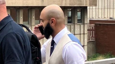Raza Mahmood, a man with a shaved head and black beard, is walking and holding a mobile phone to his ear. He is wearing a white shirt, a dark tie, and a white and blue waistcoat. In the background is Sheffield Magistrates' Court, a brutalist-style building.
